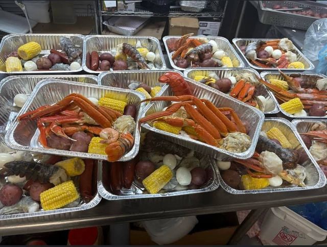 Multiple aluminum catering trays brimming with a seafood boil — bright orange crab legs and lobster tails, corn on the cob, red potatoes, boiled eggs and sausages.