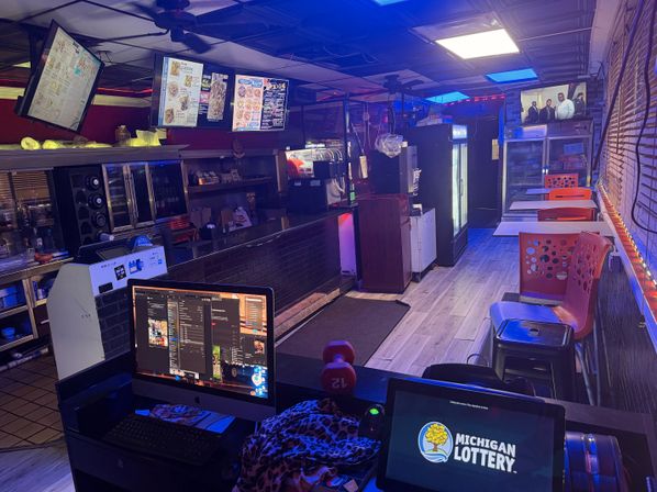 Cozy late-night takeout counter with illuminated digital menu boards above a service counter, computer monitors at the front desk, red perforated dining chairs along a window, refrigerated display cases and moody blue-purple ambient lighting.