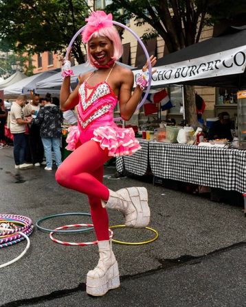 Performer in a bright pink wig and sequined tutu poses with a hula hoop at an outdoor street fair, red tights and glitter platform boots with vendor tents and extra hoops in the background.