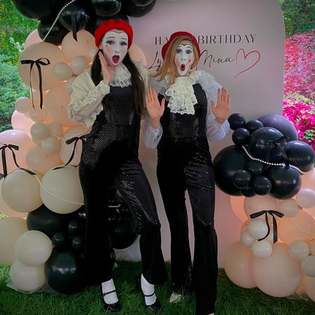 Two mimes in red berets and black sequin jumpsuits with white ruffled collars striking surprised poses in front of a blush-and-black balloon arch and birthday backdrop at an outdoor garden party
