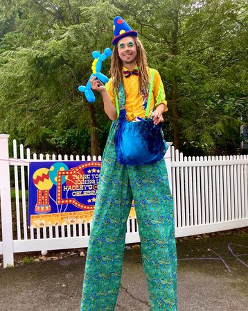 Stilt-walking clown entertainer in a polka-dot cone hat, yellow shirt, rainbow bow tie and oversized blue-green patterned pants holding a blue balloon dog at an outdoor backyard birthday celebration by a white picket fence and trees.