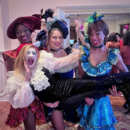 Four costumed performers in a ballroom-style party photo: three in colorful cabaret/can-can dresses with feathered headpieces holding a fourth in black sequined pants and white mime makeup, balloons and guests visible in the background.