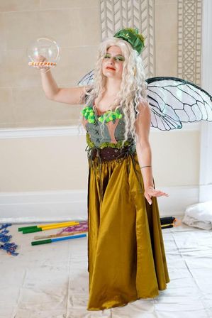 Woman in a whimsical green fairy costume with translucent wings, curly platinum hair and a gold satin skirt holding a giant soap bubble in an indoor event hall.