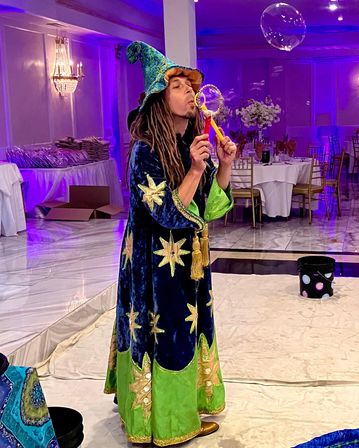 Wizard-costumed entertainer blowing a giant soap bubble in a purple-lit banquet hall with round tables, gold chairs and event decor