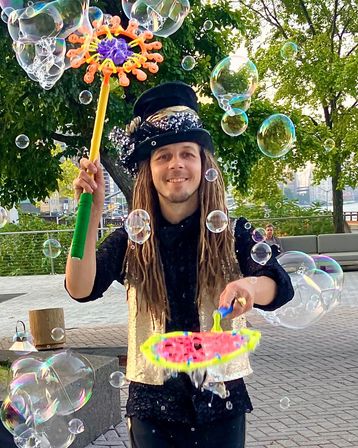 Smiling street performer in a sparkly vest and top hat creating giant iridescent soap bubbles with colorful wands at an urban waterfront park promenade