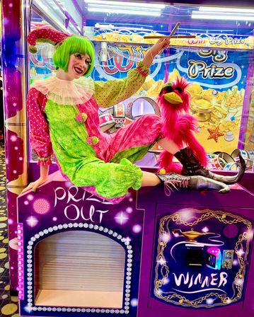 Performer in a green wig and pink-green clown costume posing on a purple arcade prize crane machine with a bright pink puppet bird, colorful lights and prize signage in the background