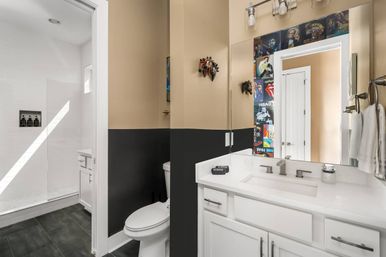 Bright modern two-tone bathroom with white vanity and rectangular sink, large mirror framed by colorful music-poster collage, glass-enclosed shower with white subway tile and built-in niche, white toilet, horse-head wall decor, dark floor tiles and towel ring with white towels.