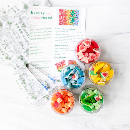 Bright flatlay of five clear jars filled with rainbow gummy candies (red, yellow, green, blue, orange) arranged around a DIY candy board instruction card and blue tongs on a white marble background