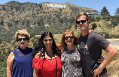Four adults smiling in sunglasses and summer outfits on a sunny trail in the Hollywood Hills, Los Angeles, with the Hollywood sign visible on the hillside behind them.
