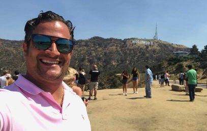 Smiling man in a pink polo and sunglasses takes a selfie at a Hollywood Hills overlook with the Hollywood Sign in the background, tourists and a sunny Los Angeles skyline