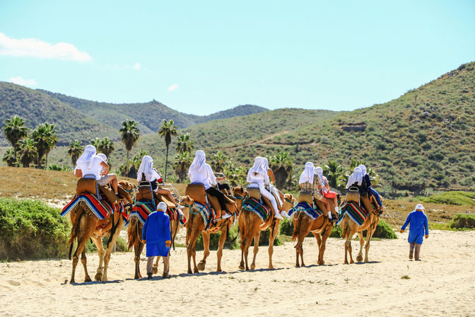 Group of tourists on a camel ride along a sandy coastal trail, riders in white headscarves seated on camels with colorful striped blankets, guides in bright blue robes walking beside them, palm trees and green hills under a clear blue sky.