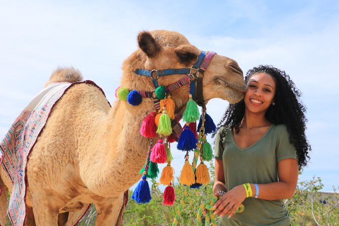 Smiling woman in a green shirt poses outdoors with a decorated camel wearing bright multicolored tassels and a patterned blanket under a sunny blue sky.