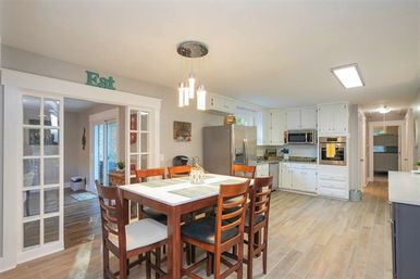 Bright open-concept kitchen and dining area with a wooden high-top table and six chairs, white cabinets, stainless steel refrigerator and built-in oven, pendant lights, wood-look tile floor, and glass-paneled doorway
