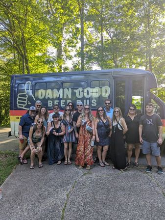 Cheery group of about 15 adults holding drinks and posing in front of a colorful beer tour bus parked on a tree-lined road on a sunny summer day.