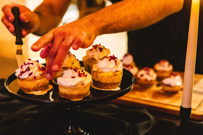 Hands arranging mini cheesecakes topped with whipped cream and dried rose petals on a black cake stand at a candlelit dessert table.