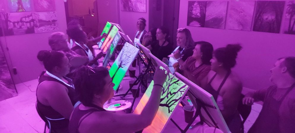 Group painting class in an indoor art studio with purple lighting, adults seated at easels painting neon green tree canvases during a fun sip-and-paint night