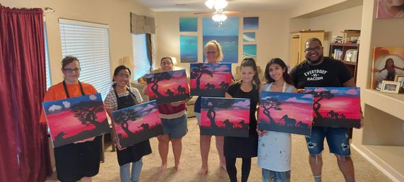 Seven people in a cozy living room posing with matching sunset-silhouette canvas paintings of acacia trees and elephants — cheerful DIY paint-night group holding finished artwork.