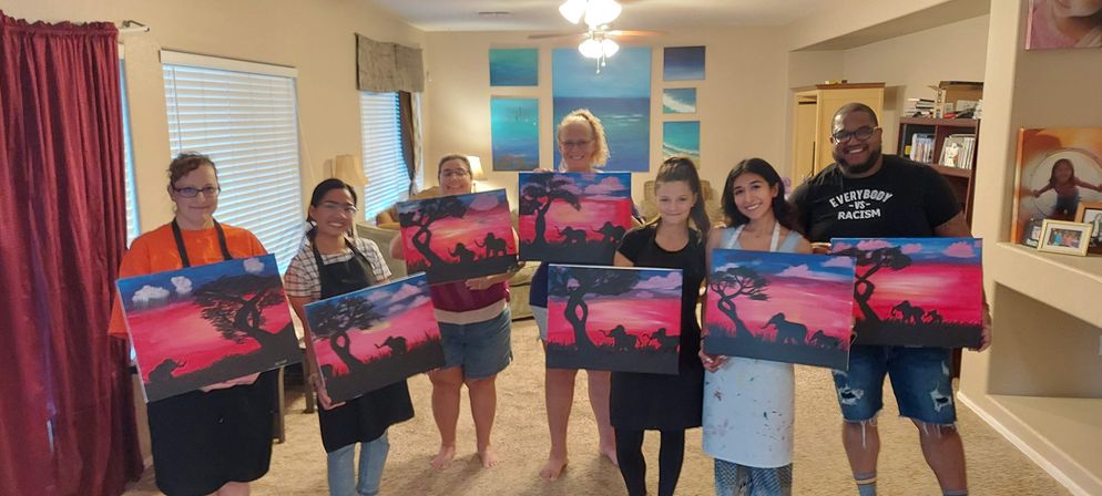Seven people in a cozy living room posing with matching sunset-silhouette canvas paintings of acacia trees and elephants — cheerful DIY paint-night group holding finished artwork.