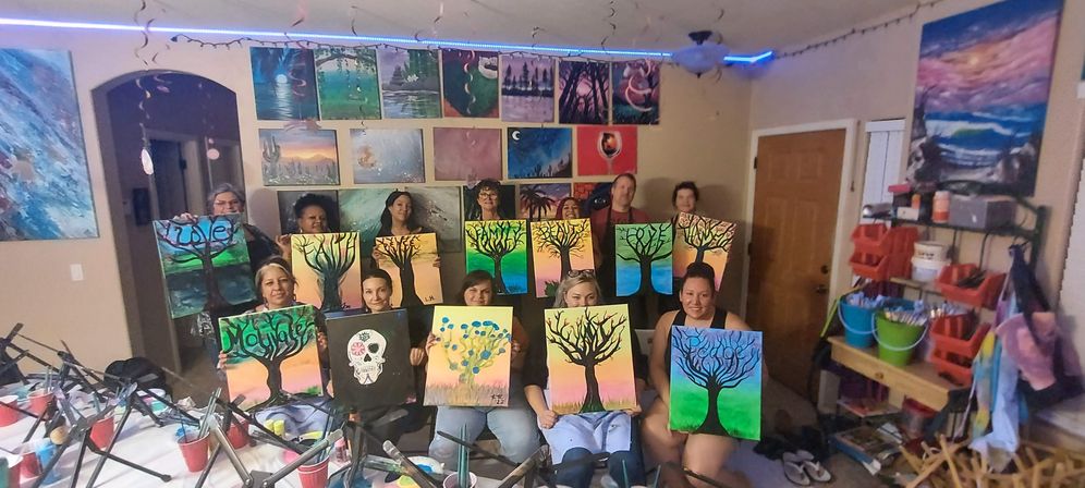 Lively indoor paint-night workshop: a group of participants in a studio proudly hold vibrant canvas tree paintings (plus one skull design), with a gallery wall and paint supplies visible around them.