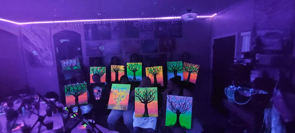 Group at an indoor glow-in-the-dark painting party holding neon tree canvases under purple LED/UV blacklight, vibrant fluorescent artwork glowing