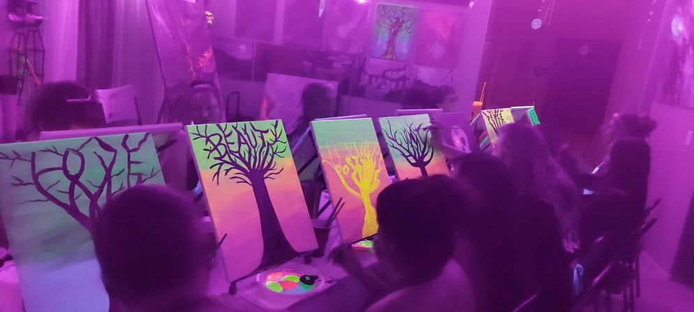 Indoor blacklight painting party in a purple-lit art studio, participants seated at easels painting neon glow-in-the-dark tree silhouette canvases.