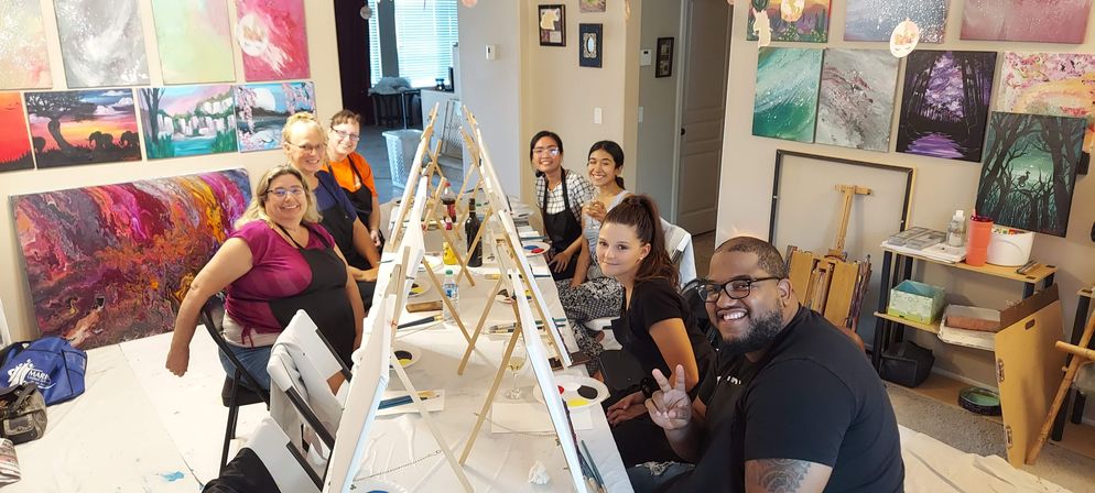 Group painting party in a bright indoor art studio: eight smiling adults seated at a long table with easels, palettes and brushes, surrounded by colorful abstract canvases on the walls.