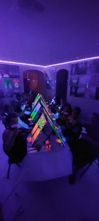 Neon blacklight painting party in an indoor studio — guests seated along a long table with back-to-back easels, glowing UV paints and vibrant canvases under purple LED strip and blacklight illumination.