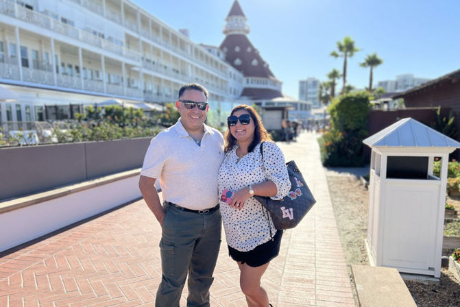 Smiling couple wearing sunglasses posing on a sunny oceanfront promenade in front of a historic Victorian-style beachfront hotel with palm trees and blue sky