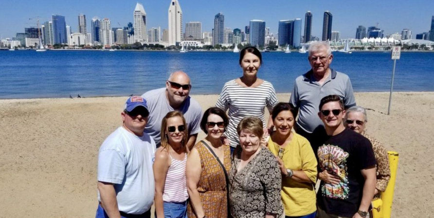 Group of adults smiling on a sunny sandy bay beach with sailboats and a modern downtown waterfront skyline across calm blue water under a clear sky.