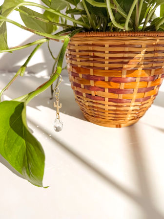 Sunlit wicker planter on a white surface with trailing pothos vine suspending a gold cactus charm and faceted teardrop crystal.