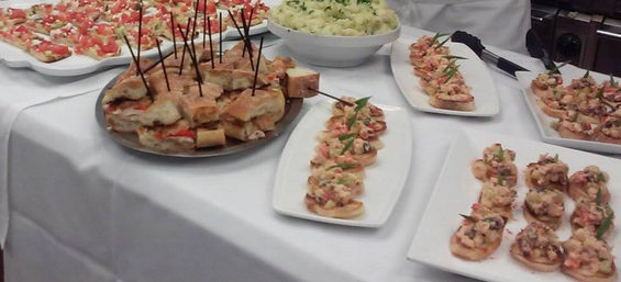 Event catering buffet on a white tablecloth with skewered mini sandwiches, seafood crostini on white platters and a bowl of creamy potato salad — tasty party hors d'oeuvres.