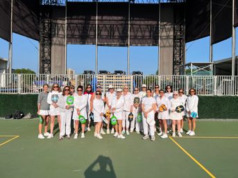 Group of women in white athletic outfits posing with colorful pickleball paddles on a sunny outdoor court in front of a large stage