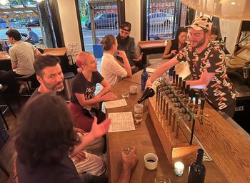 Vibrant street-front bar at night — bartender pours wine over a wooden counter with multiple taps while patrons chat over menus and drinks.