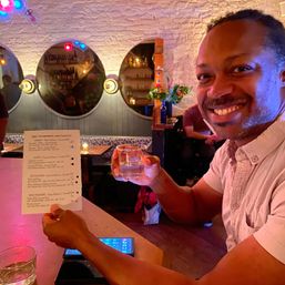 Smiling person holding a tasting glass and menu in a cozy, dimly lit bar with round mirrors, exposed white brick, colorful lights and a bottle-decorated shelf.