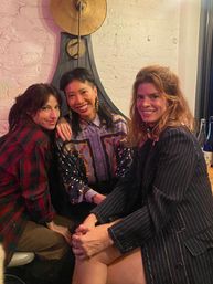 Three friends smiling and seated close in a cozy bar with exposed white brick and a large brass wall clock, wearing a patterned silk blouse, plaid shirt, and pinstripe blazer.