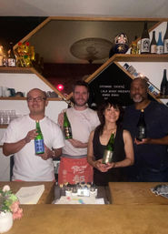 Four smiling adults behind a wooden bar holding sake bottles in a cozy Japanese‑style bar with maneki‑neko and sake bottles on hexagonal shelves.