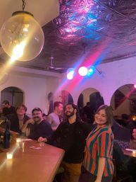 Group of friends smiling at a lively bar counter with candles and drinks, round glass pendant light, ornate tin ceiling and colorful pink-blue party lights