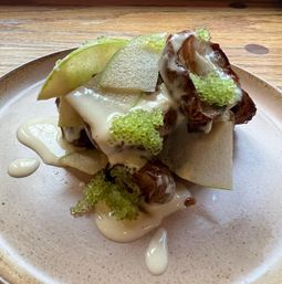 Fine-dining plated dish with thin green apple slices and crispy shards topped by caramelized brown pieces, glossy white cream and bright green caviar-like pearls on a ceramic plate.
