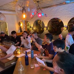 Group of people sampling drinks at a cozy craft cocktail bar counter with multiple taps, wooden bar, glowing globe pendant lights, round mirrors on exposed white brick, menus and candles creating warm ambient light.