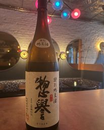 Tall Japanese sake bottle with bold calligraphy on a bar counter in a cozy, dimly lit restaurant — round mirrors, exposed white brick and colorful pendant lights overhead.