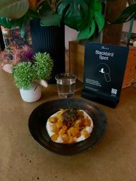 Cafe table scene with a black bowl of creamy dessert topped with diced mango and herbs, a glass of water, a small floral vase, leafy potted plant and a tabletop menu sign