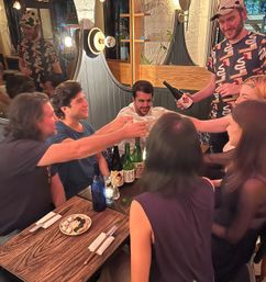 Group of friends clinking ceramic cups while a server pours sake at a cozy, dimly lit urban izakaya-style restaurant — wooden table with sake bottles, small plates and chopsticks, lively nighttime atmosphere.