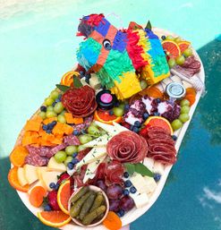 Poolside colorful piñata-topped charcuterie board with salami roses, mixed cheeses, green and red grapes, blueberries, citrus slices, dried apricots, cornichons and jam — vibrant summer party spread