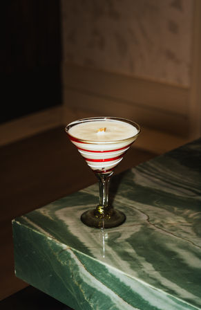 White wax candle poured into a vintage martini glass with red spiral stripes, sitting on a green marble countertop — cozy indoor tabletop decor.