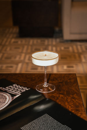 Decorative white candle poured into a gold-rimmed coupe glass on a polished wood coffee table beside an open magazine, geometric tile floor softly blurred in the background.