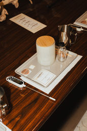 Barista toolkit on glossy dark wood table: tray with ribbed ceramic jar topped by a bamboo lid, small glass bottle, stainless steel milk pitcher, digital probe thermometer and labels — coffee brewing tools.
