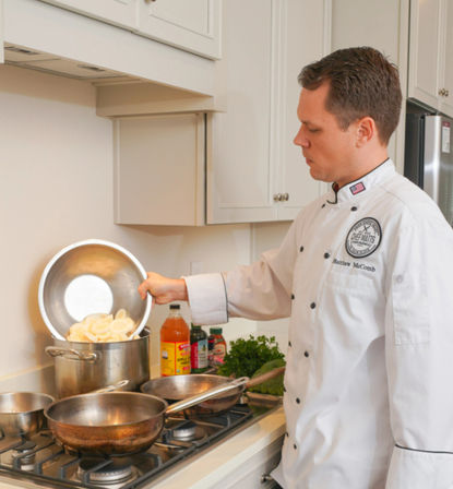 Chef in a white jacket pouring sliced potatoes from a metal bowl into a large stockpot on a gas stove, with pans, bottles and fresh herbs on a bright home kitchen counter.