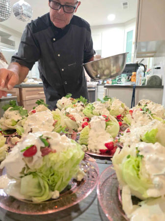 Cook in a dark jacket plating wedge salads in a bright kitchen — iceberg lettuce halves drizzled with creamy dressing, cherry tomatoes, bacon crumbles and parsley on pink plates.