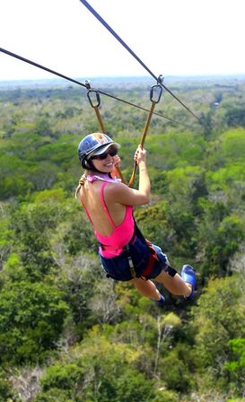 Smiling person in helmet and sunglasses ziplining over a lush green forest canopy, wearing a pink top and safety harness on an outdoor zipline adventure