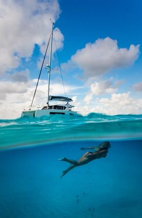 Over-under shot of turquoise tropical sea with a white catamaran on the surface and a freediver gliding underwater beneath a partly cloudy blue sky.